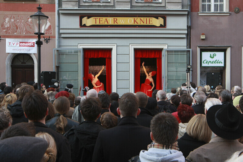 fasada kamienicy z napisem teatr w oknie, w dwóch oknach aktorzy, przed nimi widzowie