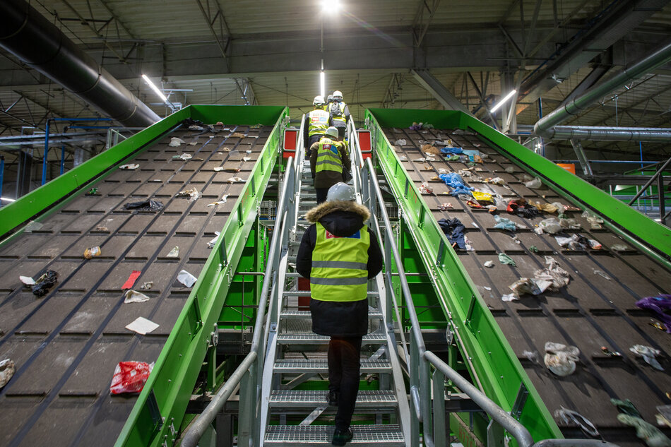 Na zdjęciu widoczne jest wnętrze obiektu przemysłowego, zakładu recyklingu lub sortowni odpadów. Kilku pracowników w odblaskowych kamizelkach i kaskach ochronnych stoi na metalowej konstrukcji z schodami. Na przenośniku rozrzucone są różne rodzaje odpadów, struktura, na której znajdują się pracownicy, jest pomalowana na zielono i wyposażona w poręcze bezpieczeństwa 