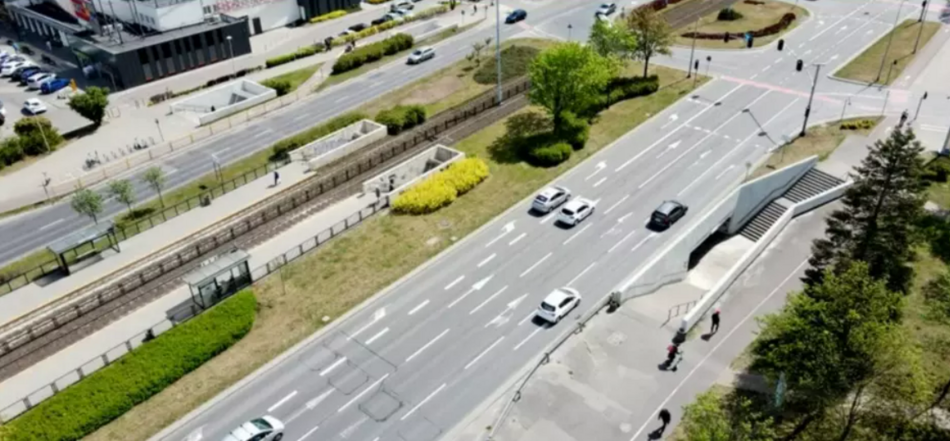 zdjęcie z drona, widać trzypasmową jezdnie na której jedzie kilak samochodów, po lewej torowisko tramwajowe, pomiędzy nimi trawnik, w tle fragment budynku