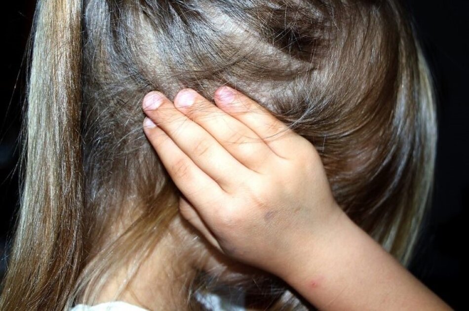 Na zdjęciu widzimy zbliżenie na głowę dziewczynki, która zakrywa uszy i twarz dłonią. Ma blond włosy, skóra dziecka jest jasna, a na małym ramieniu widoczne są drobne ślady, które mogą być naturalnymi znakami na skórze lub lekkimi zadrapaniami.