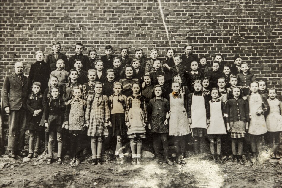 To czarno-białe zdjęcie przedstawia grupę dzieci w różnych wiekach, ustawionych w kilka rzędów, z mężczyzną, prawdopodobnie nauczycielem, po lewej stronie. Wszystkie osoby są ubrane w odzież, która wygląda na typową dla pierwszej połowy XX wieku. Dzieci stoją przed ceglaną ścianą, która może być ścianą szkoły lub innego budynku edukacyjnego. Dzieci mają poważne miny, co było charakterystyczne dla fotografii z tamtego okresu. Ubiory są stosunkowo formalne, z niektórymi chłopcami w ciemnych marynarkach lub swetrach, a dziewczętami w sukienkach i warkoczach lub kokach. Całość obrazu przekazuje wrażenie uroczystości i formalności takiego wydarzenia, jak szkolne zdjęcie grupowe