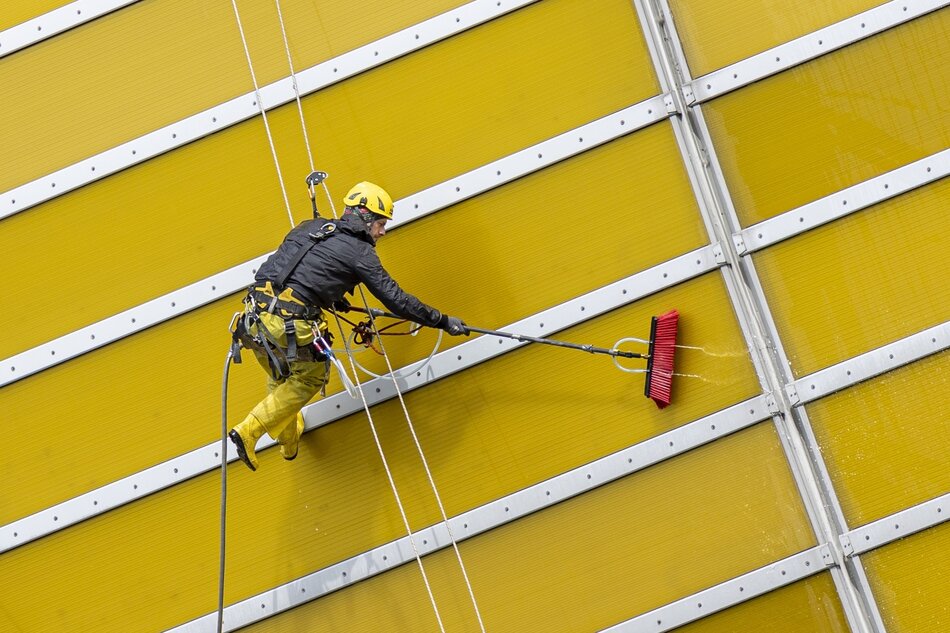 Na zdjęciu widzimy alpinistę przemysłowego pracującego na wysokości. Jest on ubrany w żółte spodnie i czarną kurtkę, a na głowie ma żółty kask, co wskazuje na stosowanie środków bezpieczeństwa. Pracownik wydaje się myć lub malować zewnętrzną ścianę budynku, używając długiego wałka z czerwoną szczotką na końcu, która jest połączona z wężem dostarczającym płyn. Jest zabezpieczony linami, które są przymocowane w wyższym punkcie konstrukcji, pozwalając na stabilną i bezpieczną pracę na wysokości. Ściana, na której pracuje, składa się z żółtych paneli podzielonych srebrnymi elementami