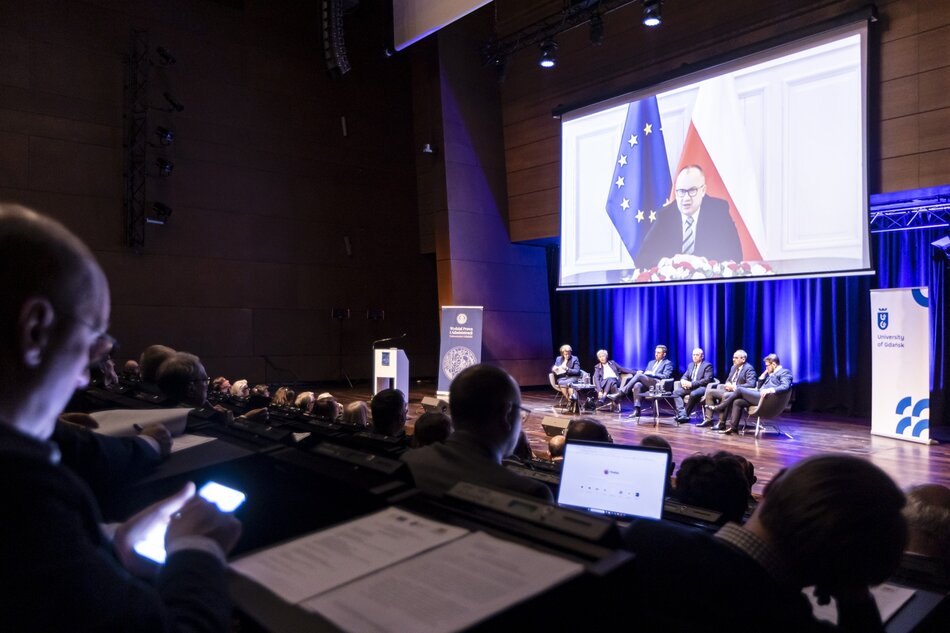 Na dużej sali konferencyjnej sześć osób siedzi na fotelach pod dużym ekranem, na którym widać przemawiającego mężczyznę. Na sali siedzą słuchacze konferencji.