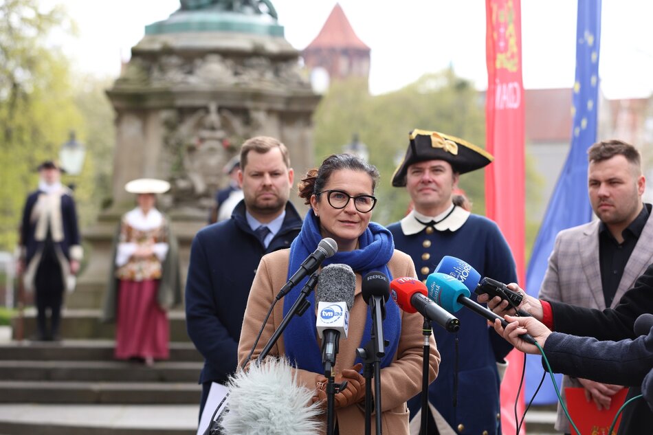Na zdjęciu widzimy kobietę stojącą przed mikrofonami, podczas konferencji prasowej. Kobieta ma na sobie szalik i okulary, a za nią widoczni są mężczyźni ubrani w historyczne kostiumy z epoki, w tym jeden w granatowym mundurze z trójgraniastym kapeluszem, który może być rekonstruktorem historycznym lub aktorem. W tle są flagi, w tym jedna Unii Europejskiej i jedna z wizerunkiem herbu, co sugeruje oficjalne wydarzenie o znaczeniu kulturalnym lub politycznym. Cała scena rozgrywa się na zewnątrz, a za uczestnikami widać pomnik i zielone otoczenie.