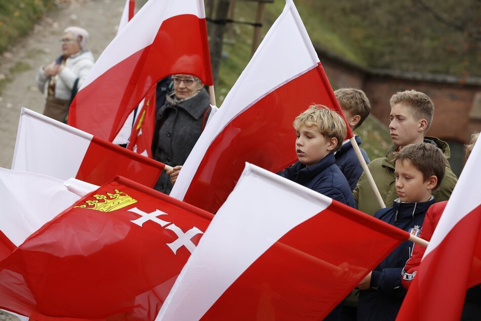 Na zdjęciu widzimy grupę dzieci niosących flagi. Flaga, którą niosą, to prawdopodobnie flaga Polski z dodatkowym godłem w centrum – białym orłem na czerwonym tle. Dzieci mają na sobie zimową odzież, co sugeruje chłodniejszą pogodę. W tle dostrzec można starszych ludzi obserwujących lub towarzyszących dzieciom, co może wskazywać na jakiś oficjalny marsz, paradę lub inną ceremonię. Otoczenie jest nieco nieostre, ale można zauważyć, że wydarzenie to odbywa się na zewnątrz, być może w historycznym lub ważnym dla społeczności miejscu.