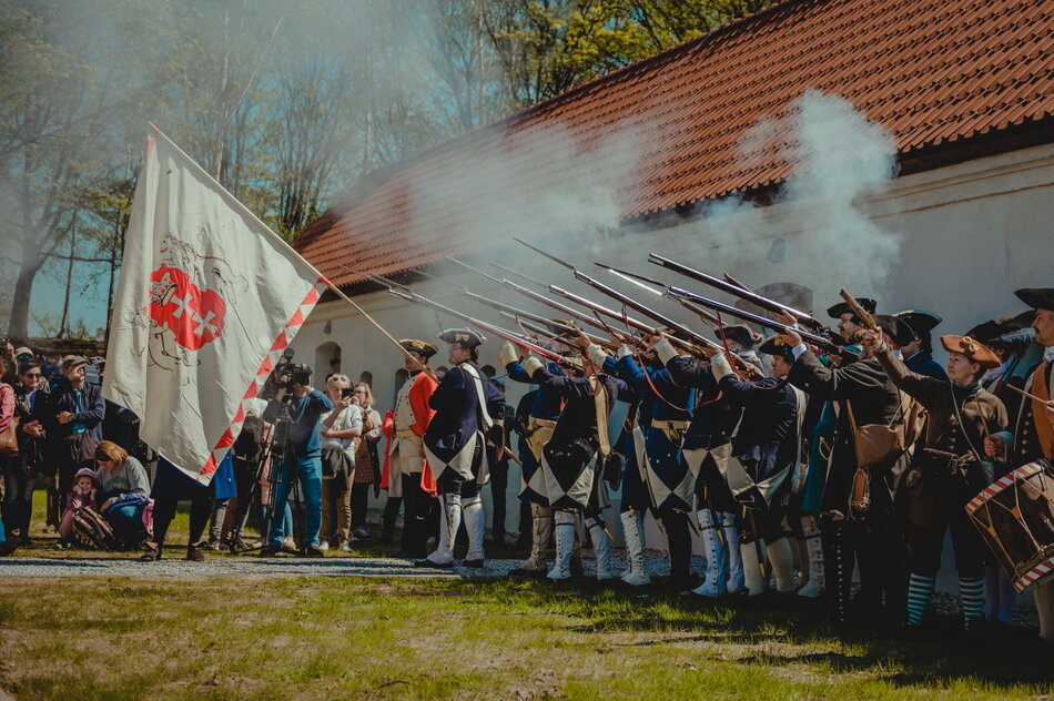 Mężczyźni w mundurach z epoki napoleońskiej, którzy są rekonstruktorami oddają salwę honorową z muszkietów. Jeden z żołnierzy trzyma flagę z herbem Gdańska. W tle odrestaurowany budynek koszarów.