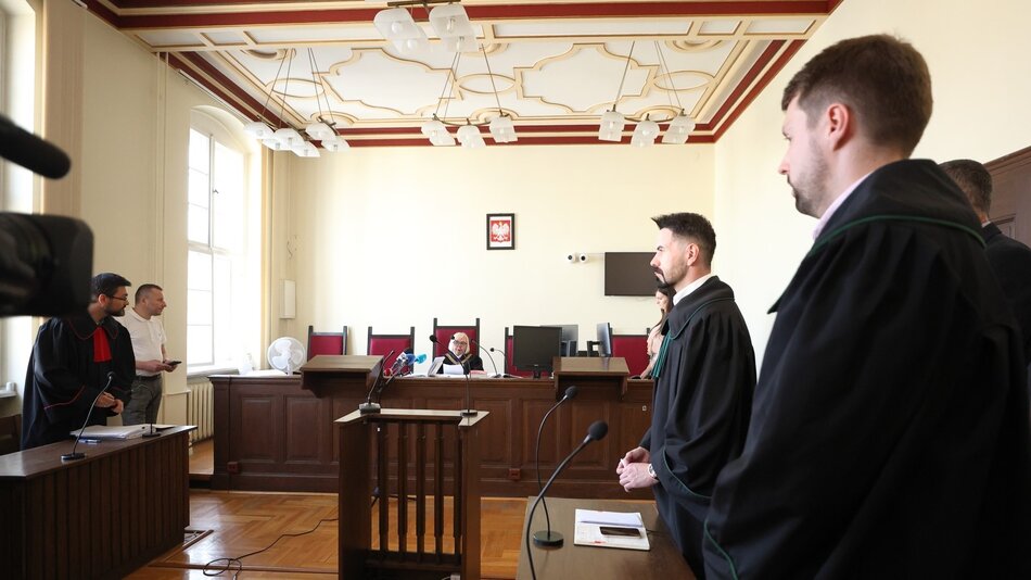 Na zdjęciu widoczna jest sala sądowa. W centralnej części znajduje się mężczyzna stojący przodem do sędziego, który siedzi za pulpitem w tle. Mężczyzna ma ciemne włosy i jest ubrany w czarną togę adwokacką, co sugeruje, że może być adwokatem lub prokuratorem. Po jego lewej stronie stoi inny mężczyzna w białej koszulce i spodniach, również skierowany w stronę sędziego, co może wskazywać, że jest stroną w sprawie lub świadkiem. Po prawej stronie stoi kolejna osoba w togi adwokackiej. W tle po prawej stronie znajdują się dwie osoby siedzące, które mogą być stronami procesu lub ich reprezentantami. Całość jest obserwowana przez kamerę filmową, co wskazuje na to, że postępowanie może być rejestrowane lub transmitowane. Sala ma klasyczny wystrój, z wysokimi oknami i ozdobnym sufitem. Na ścianie wisi herb, prawdopodobnie narodowy.