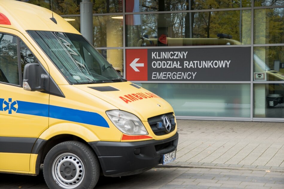 Z lewej strony stoi żółty ambulans stojący przed wejściem do szpitalnego oddziału ratunkowego. Pojazd jest wyposażony w niebieskie pasy i oznaczenia z symbolem medycznym. Znajduje się przed szklanym wejściem do budynku, na którym znajduje się duży napis Kliniczny Oddział Ratunkowy oraz angielski odpowiednik Emergency. W tle widać drzewa, co sugeruje, że szpital znajduje się w spokojnej, zielonej okolicy.