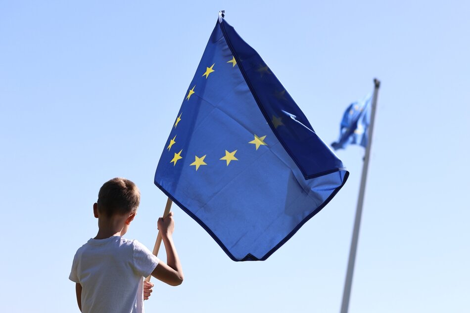 Na zdjęciu widać młodego chłopca trzymającego flagę Unii Europejskiej. Chłopiec stoi na tle błękitnego nieba, a flaga jest wyraźnie widoczna z dwunastoma złotymi gwiazdami rozmieszczonymi w kółku na niebieskim tle. W tle nieco rozmyta widać także inną flagę UE na maszcie. 