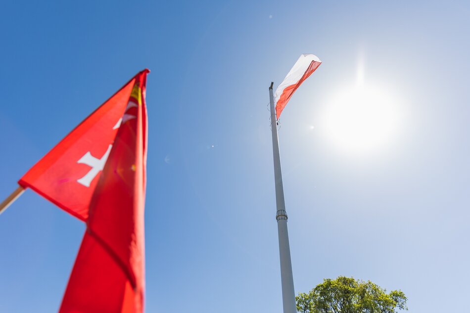 Na pierwszym planie po lewo fragment czerwonej flagi z godłem Gdańska. W tle wysoki maszt a na nim biało-czerwona flaga