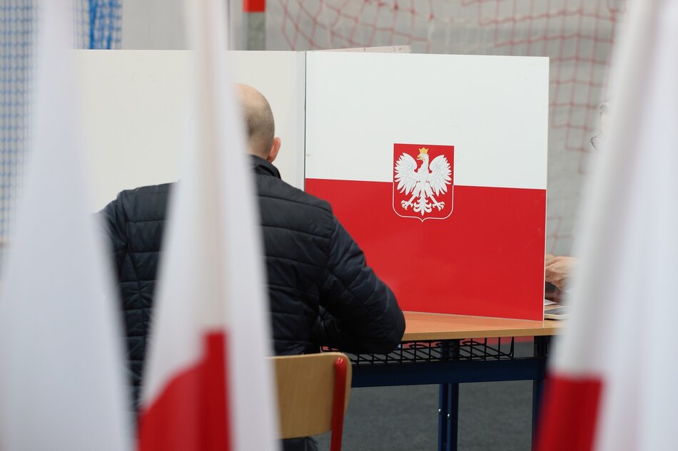 Na zdjęciu widać wnętrze jasnego pomieszczenia, prawdopodobnie szkoły lub innego budynku publicznego, gdzie odbywa się proces głosowania. Mężczyzna wkłada swój głos do przezroczystej urny wyborczej. Obok niego stoją dwoje małych dzieci, które również trzymają w rękach karty do głosowania, być może pomagając dorosłym. W tle widoczne są polskie flagi oraz kabiny do głosowania z emblematem Polski na białych parawanach. Cała scena wydaje się być momentem zaangażowania obywatelskiego, z udziałem różnych pokoleń. Atmosfera jest spokojna i oficjalna.
