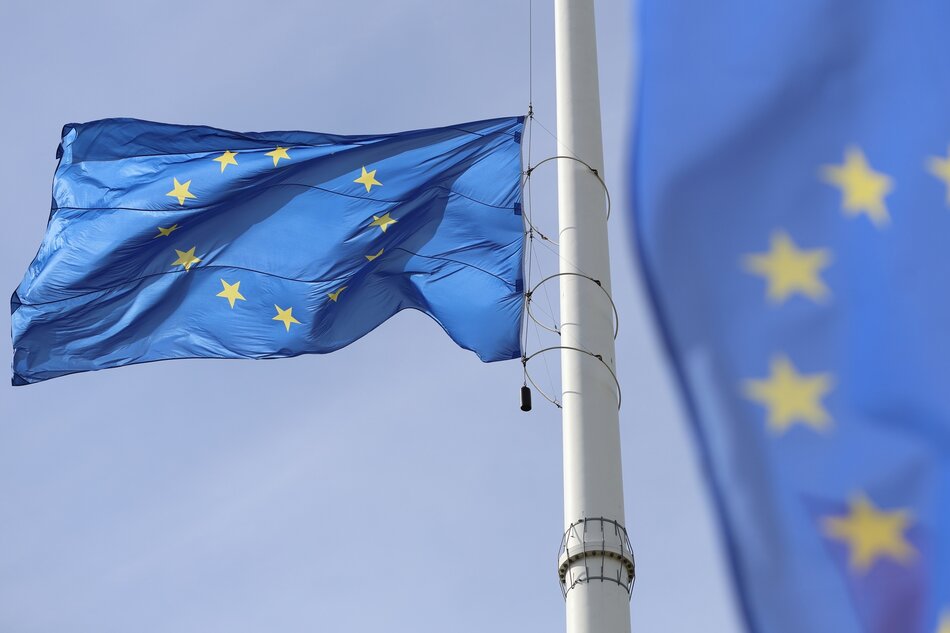 Na zdjęciu widoczna jest flaga Unii Europejskiej powiewająca na wietrze. Flaga jest koloru niebieskiego z kołem dwunastu złotych gwiazd w centrum. Tło za flagą jest jasne, co sugeruje pogodny dzień. Flagę utrzymuje biały maszt, który jest wyposażony w liny i metalowe okucia, pomagające utrzymać flagę na miejscu. Flaga zdaje się być w dobrym stanie, z wyraźnymi, nienaruszonymi kolorami i kształtem.