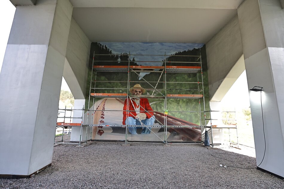 Mural przedstawiający mężczyznę w jasnym kapeluszu, czerwonej marynarce i błękitnych dżinsach, który siedzi na torach kolejowych. Malunek zdobi ściany pod wiaduktem kolejowym. Przysłania go częściowo rusztowanie.