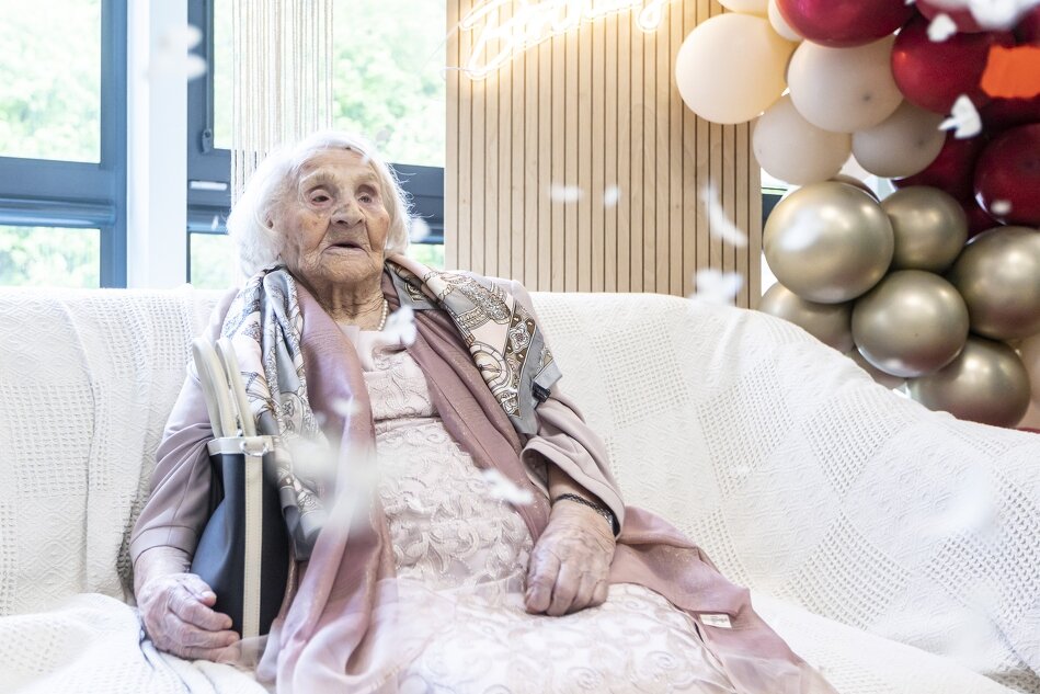 Na zdjęciu widzimy starszą panią, siedzącą wygodnie na białej sofie, ubraną w elegancką, pastelową suknię i szalik. W tle rozciągają się kolorowe balony, dodając celebracyjnego nastroju do tej wyjątkowej okazji