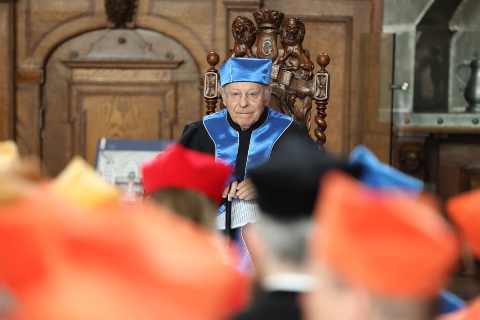 Na zdjęciu widzimy starszego mężczyznę w niebieskiej akademickiej todze, stojącego za mównicą w eleganckiej drewnianej sali. Jego poważna postawa i akademicki strój wskazują na jego uczestnictwo w ważnym wydarzeniu uniwersyteckim 