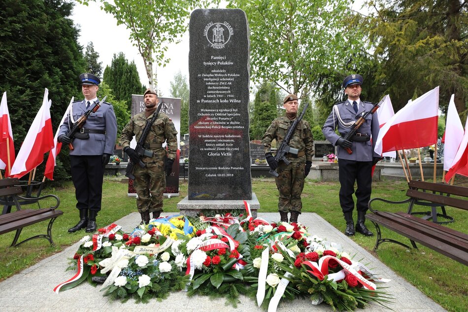 na zdjęciu pomnik w postaci wysokiego ciemnego głazu, stoją przy nim na baczność policjanci i żołnierze w oficjalnych mundurach