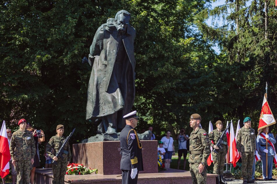 Na zdjęciu widzimy ceremonię wojskową przy pomniku z brązu przedstawiającym dwie postacie, prawdopodobnie ważne historyczne osobistości. Osoby w mundurach wojskowych, w tym oficer w czapce i zdejmujący okulary, stoją na tle zielonych drzew, a tło sceny zdobią flagi narodowe.