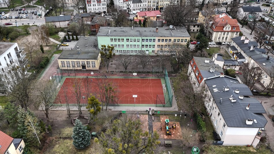 zdjęcie z drona, widać szkolne boisko o bordowej nawierzchni, wokół niego ustawione są prostokątne budynki szkolne