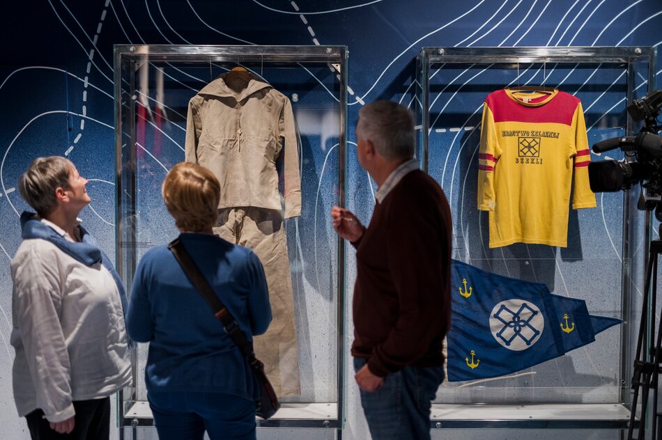 Na zdjęciu trzy osoby w średnim wieku, dwie kobiety i mężczyzna, oglądają eksponaty w muzeum, zainteresowane historycznymi artefaktami związanymi z morzem. W szklanych gablotach wyeksponowane są przedmioty takie jak stary mundur żeglarski i koszulka z żeglarskimi insygniami, co podkreśla morski temat wystawy.