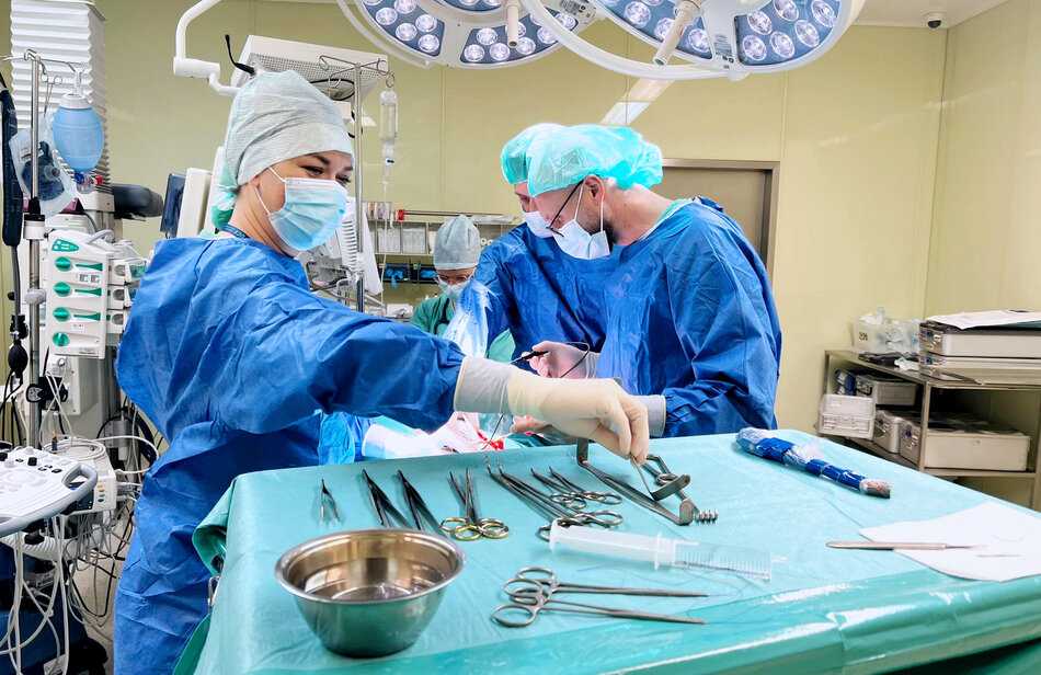 Na zdjęciu widzimy zespół chirurgiczny w trakcie operacji w szpitalnej sali operacyjnej. Personel w niebieskich uniformach i maskach skupia się na zabiegu, korzystając z różnych narzędzi chirurgicznych rozmieszczonych na zielonym stole operacyjnym.