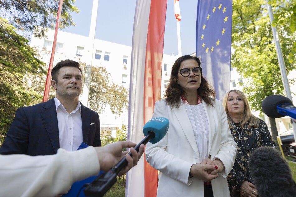 Trzy osoby, dwie kobiety i mężczyzna stoją przed mikrofonami podczas konferencji prasowej. Za nimi flagi Polski i Unii Europejskiej. W tle budynek Urzędu Miasta.