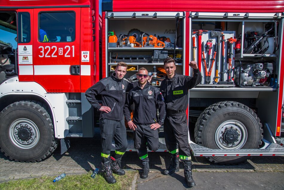 Na zdjęciu trzech strażaków stoi dumnie przed wozem strażackim. Są ubrani w standardowe, czarne uniformy z odblaskowymi elementami, a za nimi widać otwarty przedział z narzędziami ratunkowymi i sprzętem pożarniczym.