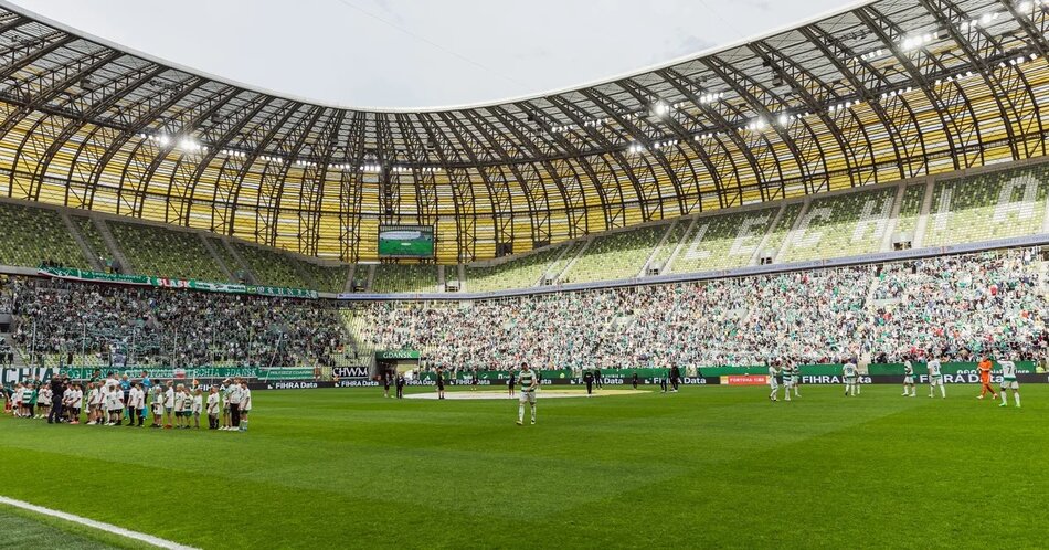 Zdjęcie przedstawia nowoczesny stadion piłkarski w trakcie meczu, pełen kibiców ubranych głównie w biało-zielone barwy. Stadion charakteryzuje się imponującą, zakrzywioną, żółto-zieloną konstrukcją dachu, która tworzy efektowną oprawę dla sportowych wydarzeń.