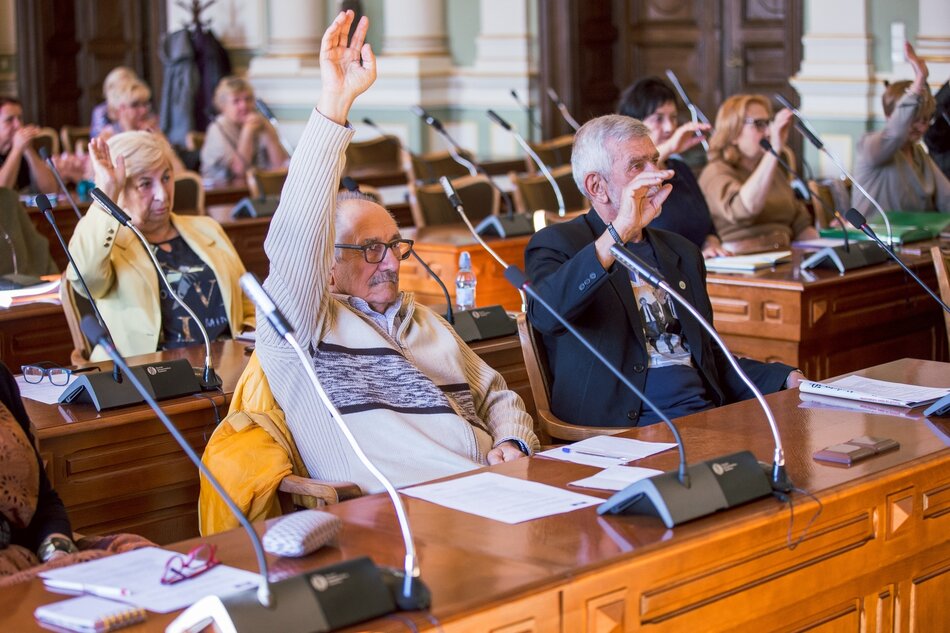 na zdjęciu kilka starszych osób siedzi za dużymi drewnianymi stołami, na pierwszym planie dwóch starszych mężczyzn podnosi rękę w geście głosowania