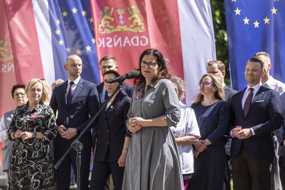 Zdjęcie przedstawia grupę pięciu osób stojących na zewnątrz, uśmiechających się do kamery. W tle widoczne są kolorowe flagi, między innymi czerwone, niebieskie i białe, co sugeruje, że może to być wydarzenie oficjalne lub patriotyczne. Osoby na zdjęciu są elegancko ubrane, większość w garnitury lub marynarki. Trzy kobiety i dwóch mężczyzn stoją w rzędzie obok siebie. Kobieta w środku nosi szarą sukienkę z paskiem, natomiast pozostałe osoby mają na sobie ciemne, formalne ubrania. Każda z osób jest dobrze wystylizowana, co może wskazywać na ich zawodowe lub oficjalne role. W tle, za osobami, widoczne są również drzewa i budynki, co sugeruje, że zdjęcie zostało zrobione na świeżym powietrzu w mieście lub na terenie instytucji.