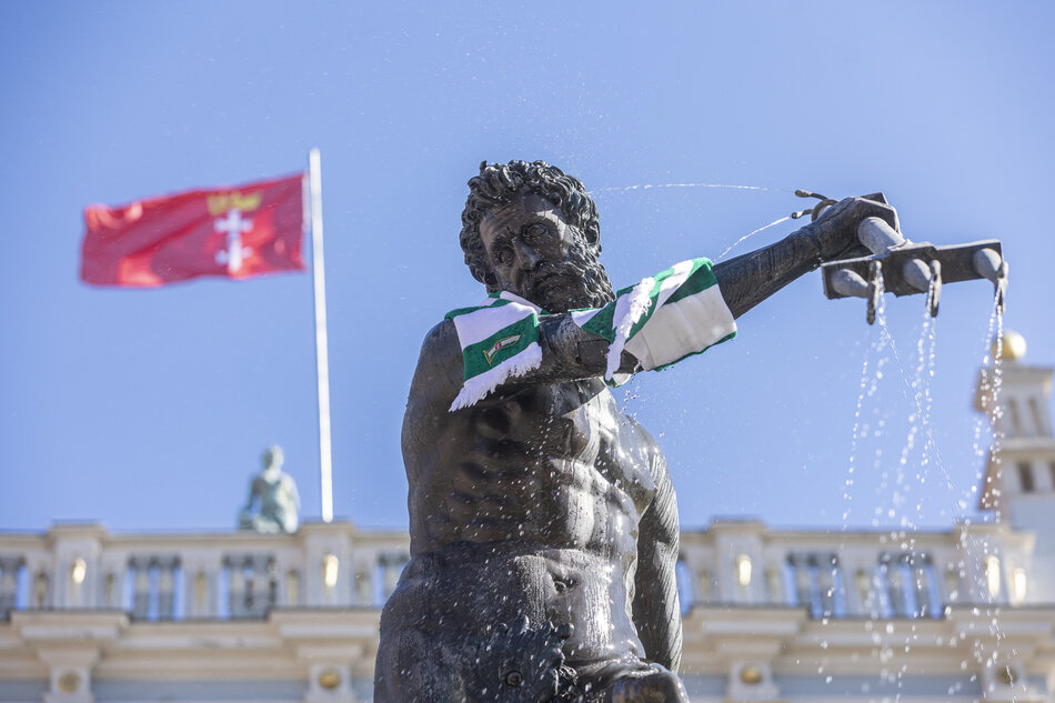 metalowa figura przedstawiająca brodatego mężczyznę owinięta wełnianym szalikiem. z tyłu widoczny zabytkowy budynek, na szczycie którego powiewa flaga