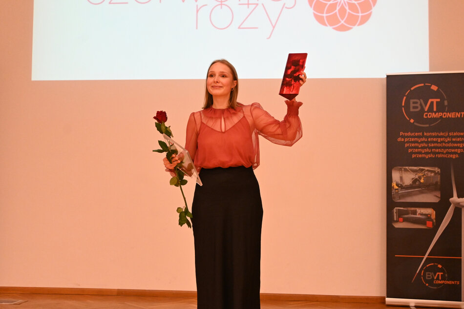 Młoda kobieta, studentka stoi uśmiechnięta na scenie. W jednym ręku trzyma kwiat - czerwoną różę, w drugim ręku trzyma statuetkę Czerwonej Róży - nagrodę w konkursie. 