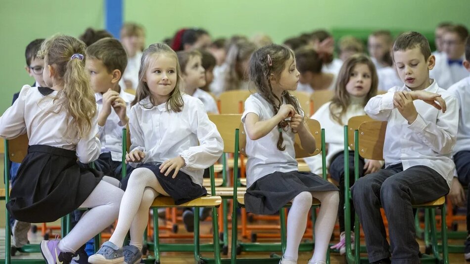 Zdjęcie przedstawia grupę dzieci siedzących na krzesłach w ustawieniu klasowym lub szkolnym. Dzieci są ubrane odświętnie, w białe koszule i ciemne spódnice lub spodnie. W tle widać więcej dzieci w podobnym stroju, sugerując, że może to być jakaś szkolna uroczystość lub przedstawienie. Na pierwszym planie znajdują się trzy dziewczynki i jeden chłopiec, którzy zdają się być zajęci rozmową lub czymś innym, czekając na rozpoczęcie wydarzenia.