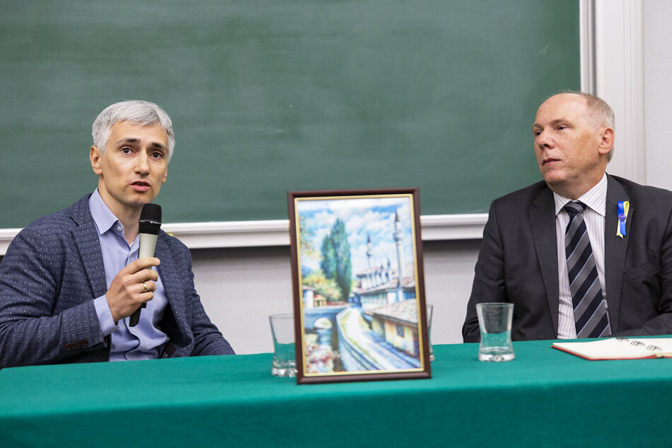 Dwóch mężczyzn siedzi za stołem z mikrofonami. Pomiędzy nimi stoi obrazek z widokiem miasta na Krymie.