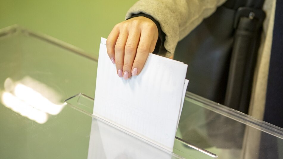 Zdjęcie przedstawia zbliżenie ręki osoby wrzucającej kartę do głosowania do przezroczystej urny wyborczej. Ręka ma zadbane, jasnoróżowe paznokcie. Osoba ta ubrana jest w jasną kurtkę. Tło zdjęcia jest nieostre, co sprawia, że głównym punktem ostrości jest ręka i karta wyborcza.