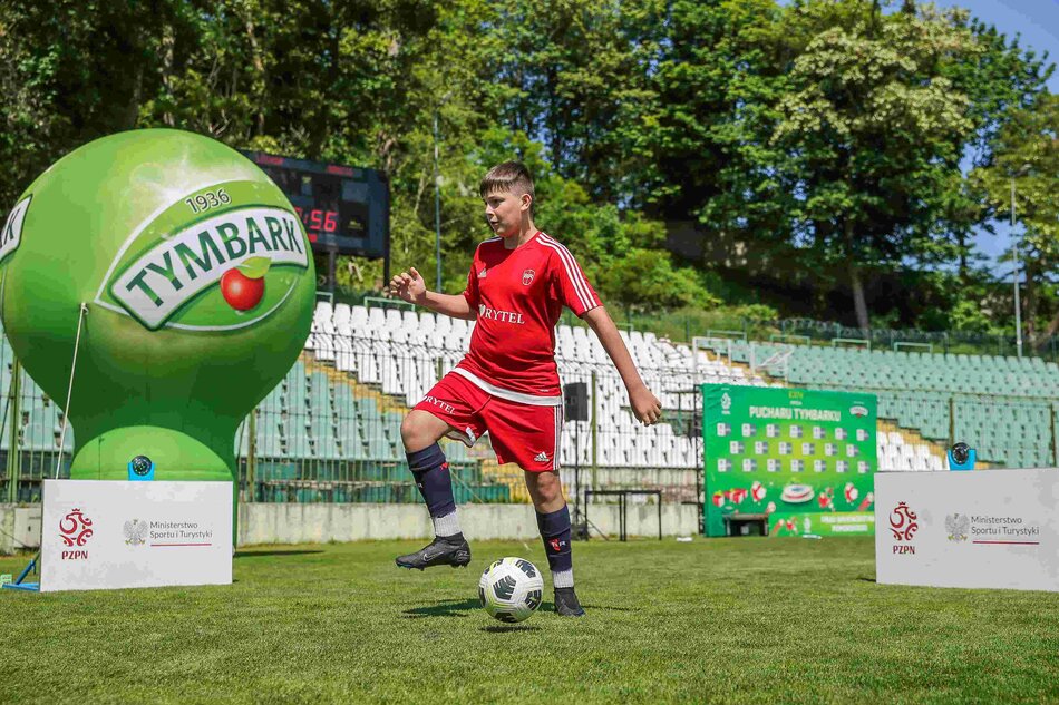 Młody piłkarz z piłką na boisku. Po lewej duży balon z napisem Puchar Tymbarku