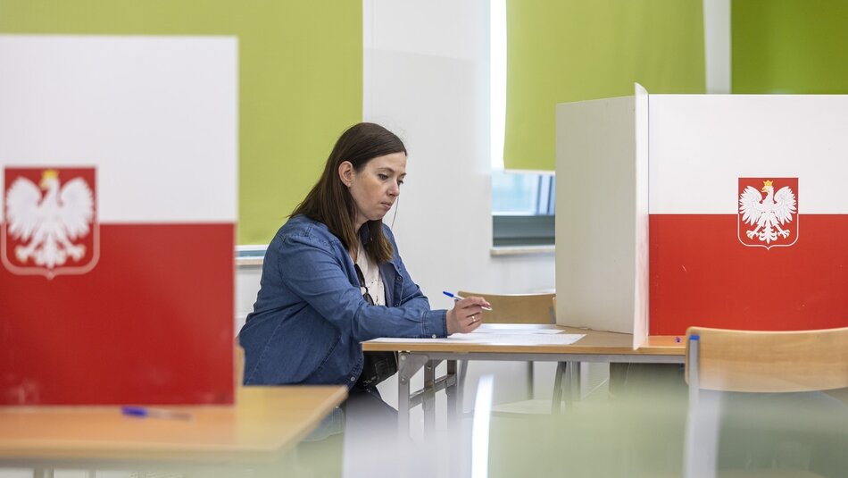 Zdjęcie przedstawia kobietę siedzącą przy stole w lokalu wyborczym, w Polsce. Kobieta jest ubrana w niebieską dżinsową koszulę i trzyma długopis, prawdopodobnie wypełniając kartę do głosowania. Przed nią znajduje się biała przegroda z godłem Polski, czyli białym orłem na czerwonym tle. W tle widać jasne ściany i zielone zasłony. Na stole obok przegrody leży kolejny długopis. Atmosfera na zdjęciu sugeruje, że kobieta jest skupiona na swojej czynności.