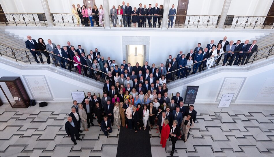 Grupa ludzi zgromadzona na reprezentacyjnych schodach w eleganckim budynku, prawdopodobnie podczas ważnego spotkania lub konferencji. Uczestnicy są ubrani formalnie, co sugeruje oficjalny charakter wydarzenia.