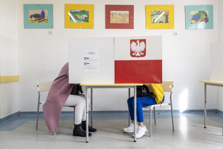 na zdjęciu stolik na którym ustawiono duża tablicę, zasłaniającą dwie głosujące osoby, obie siedzą, widać tylko ich nogi, na tablicy z jej prawej strony jest namalowana biało czerwona flaga