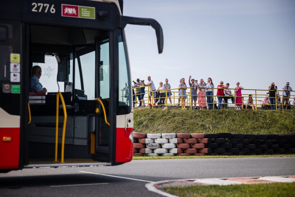 Na pierwszym planie nieostry fragment autobusu w ruchu po torze, przez otwarte drzwi widać kierowcę. W tle na wale ziemnym otoczonym oponami stoi publiczność. Ludzie śmieją się i machają rękoma. 