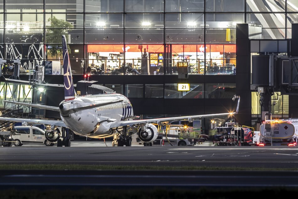 Zdjęcie przedstawia samolot pasażerski przygotowywany do lotu na płycie lotniska w nocy, z terminalem pasażerskim i światłami wewnątrz budynku w tle.