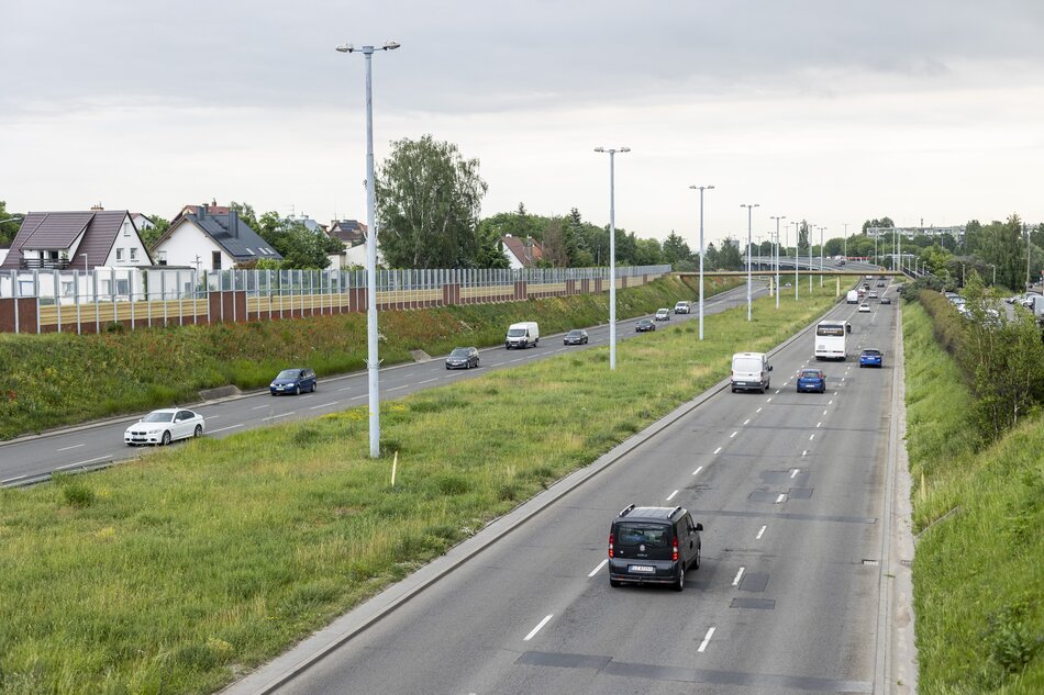 Zdjęcie przedstawia dwupasmową drogę, po której poruszają się samochody osobowe i dostawcze w obu kierunkach. Wzdłuż drogi znajdują się latarnie uliczne oraz ekrany dźwiękochłonne oddzielające jezdnię od pobliskich domów jednorodzinnych.