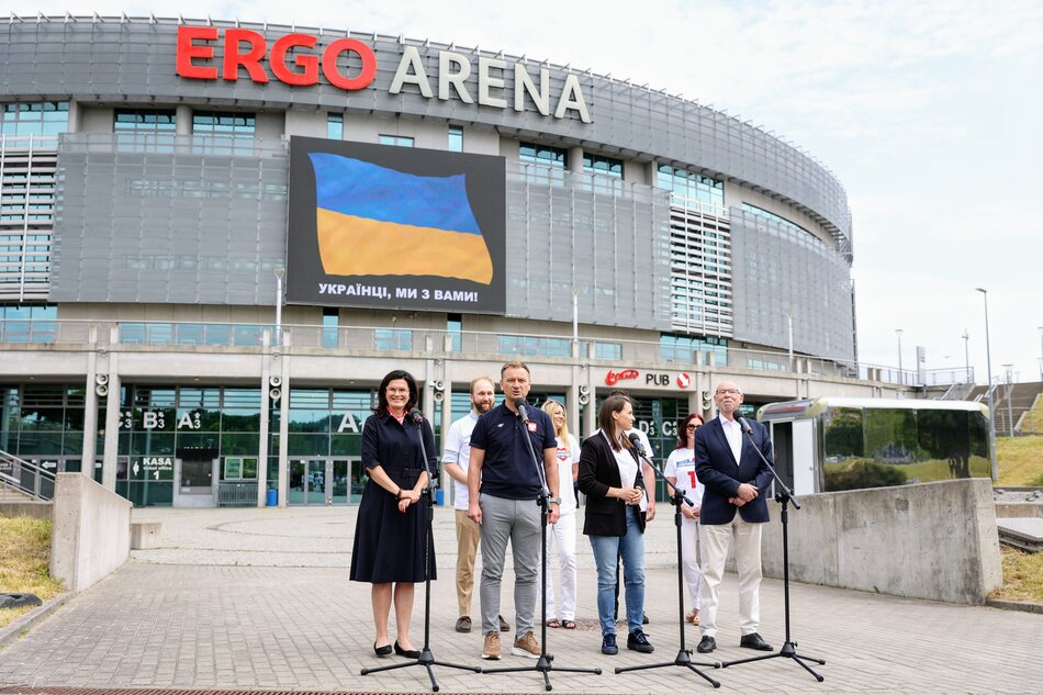 Czworo osób, dwie kobiety i dwóch mężczyzn stoi przed mikrofonami podczas konferencji prasowej zaaranżowanej przed hala sportową. Za nimi stoi jeszcze kilka osób. W tle widać halę sportową z napisem Ergo Arena.