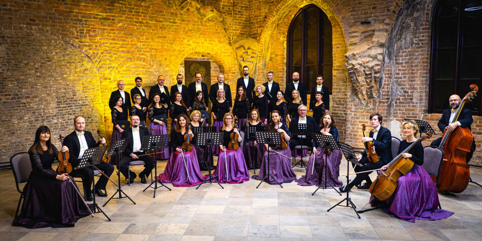 Zdjęcie przedstawia orkiestrę kameralną i chór Cappella Gedanensis, ustawioną na tle ceglanej ściany w historycznym wnętrzu. Muzycy i śpiewacy ubrani są elegancko, kobiety w fioletowych sukniach, a mężczyźni w czarnych garniturach z muszkami, co tworzy formalną i uroczystą atmosferę