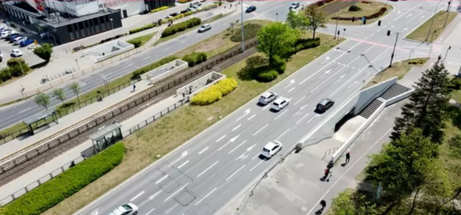 zdjęcie z drona, widać trzypasmową ulicę po której jedzie kilka samochodów osobowych, po lewej fragment torowiska i przejścia podziemnego