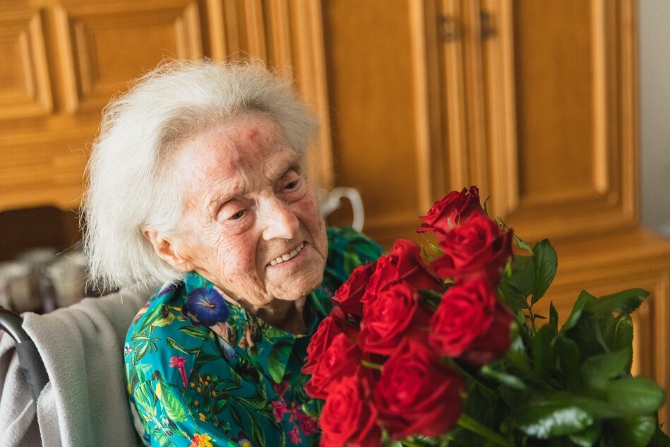 Na zdjęciu widzimy starszą kobietę z siwymi włosami, która trzyma bukiet czerwonych róż i uśmiecha się delikatnie. Kobieta ma na sobie kolorową bluzkę w kwiaty, a w tle widoczne są drewniane szafki.