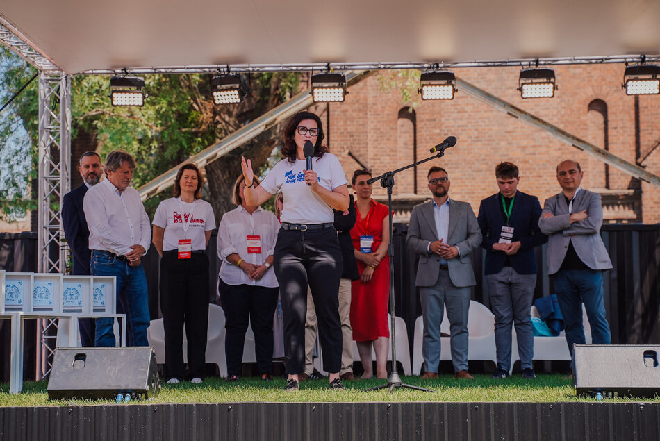 Kilka osób odświętnie ubranych na scenie. W środku kobieta z mikrofonem