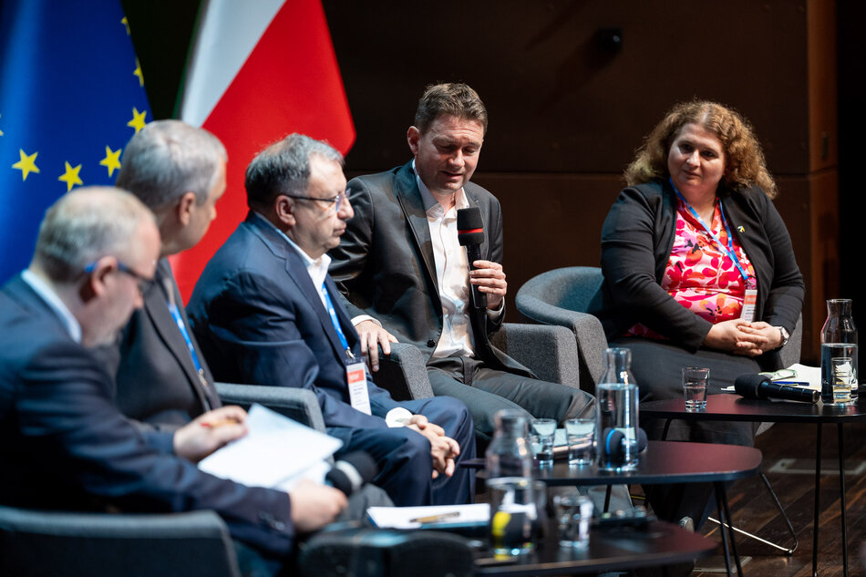 Na zdjęciu widać pięciu uczestników panelu dyskusyjnego siedzących w półokręgu na scenie, z czego jeden mężczyzna trzyma mikrofon i przemawia, a pozostali słuchają. W tle widoczne są flagi Unii Europejskiej i Polski, a na stoliku przed panelistami znajdują się butelki z wodą i szklanki