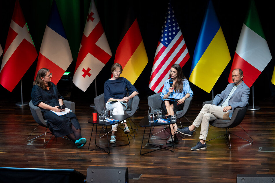 Na zdjęciu widzimy cztery osoby siedzące na scenie w fotelach, z mikrofonami w rękach, uczestniczące w panelu dyskusyjnym. W tle znajdują się flagi różnych krajów, w tym Danii, Francji, Gruzji, Niemiec, Stanów Zjednoczonych, Ukrainy i Włoch.