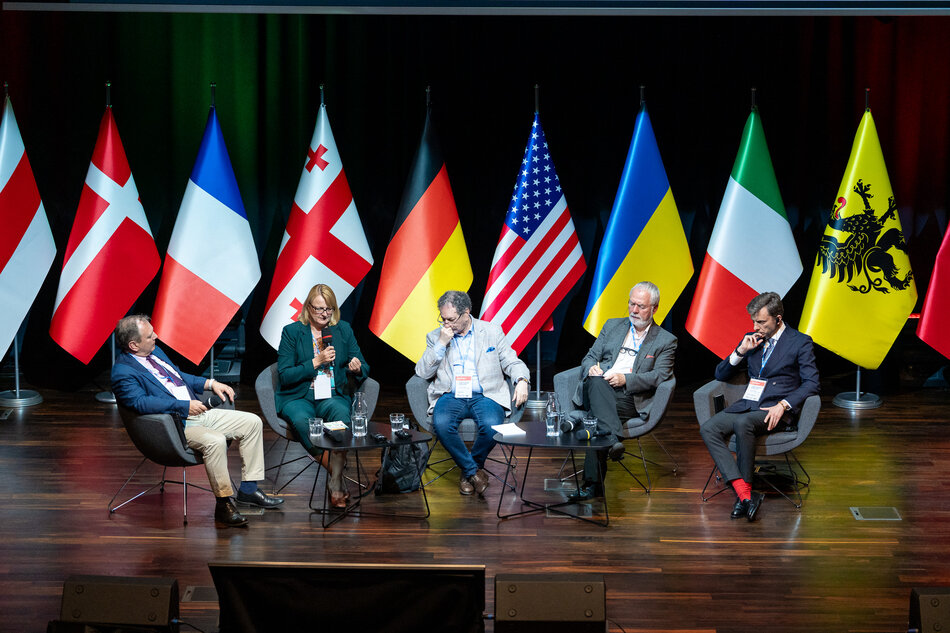 Na zdjęciu widzimy panel dyskusyjny z udziałem pięciu osób, które siedzą na scenie przed flagami różnych krajów, w tym Francji, Niemiec, Stanów Zjednoczonych, Ukrainy, Włoch i Belgii. Uczestnicy są zaangażowani w rozmowę, a jeden z nich, kobieta w zielonym garniturze, trzyma mikrofon i przemawia do publiczności.