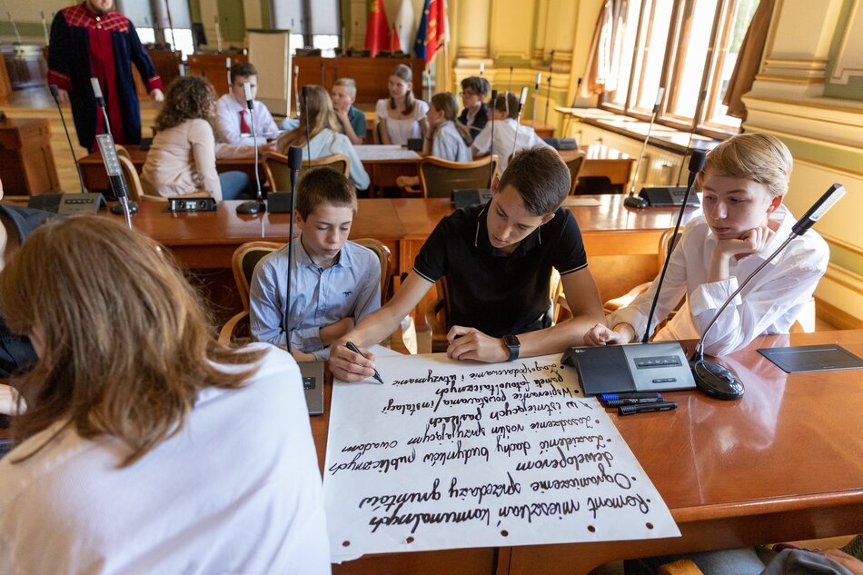 na zdjęciu kilkoro uczniów siedzi przy dużym brązowym stole, pisze coś flamastrem na ogromnej białej kartce